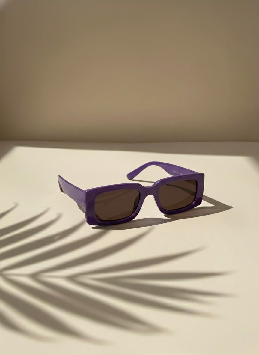 Bold purple rectangular sunglasses with smoky lenses resting on a sunlit surface with palm shadows — VA RA Blackberry Martini Sunglasses.