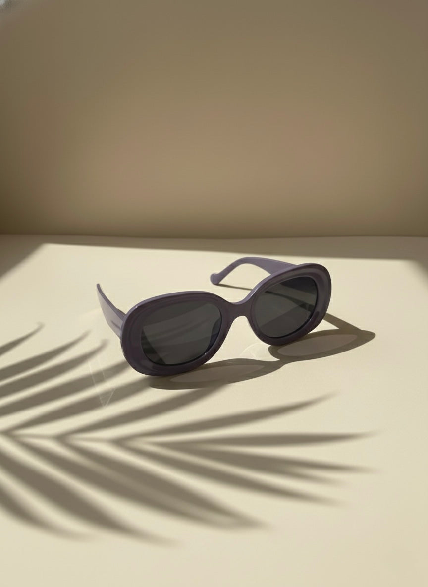 Purple oval sunglasses resting on a sunlit surface with palm shadows — VA RA Grape Crush Sunglasses.