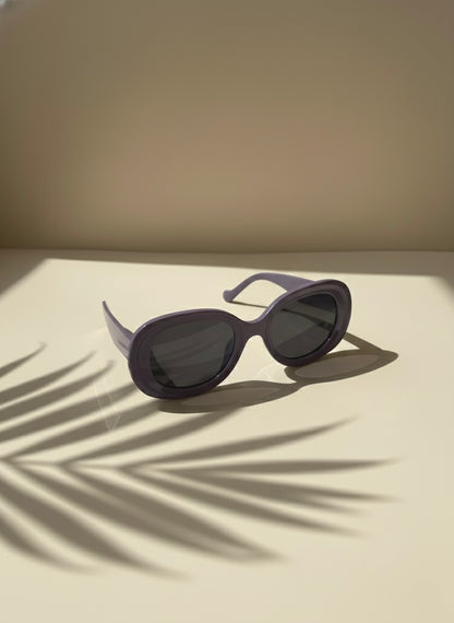 Purple oval sunglasses resting on a sunlit surface with palm shadows — VA RA Grape Crush Sunglasses.