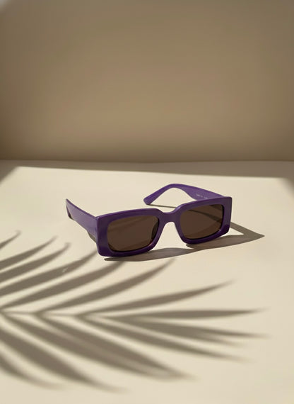 Bold purple rectangular sunglasses with smoky lenses resting on a sunlit surface with palm shadows — VA RA Blackberry Martini Sunglasses.